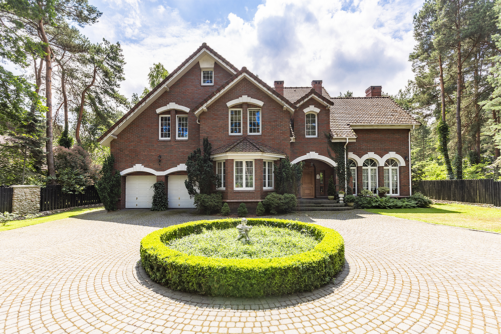 Elegant red brick house with plantation shutters, circular landscaped garden, and driveway, surrounded by trees, emphasizing home aesthetics and energy efficiency.