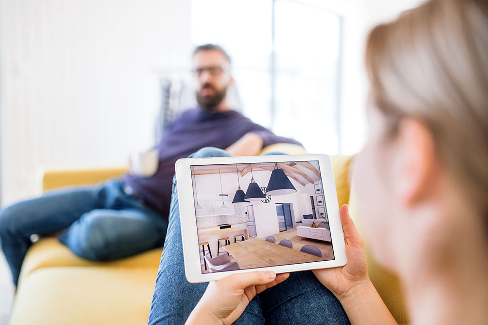 Couple reviewing home design on tablet while seated on a sofa, showcasing modern interior with plantation shutters and stylish decor.