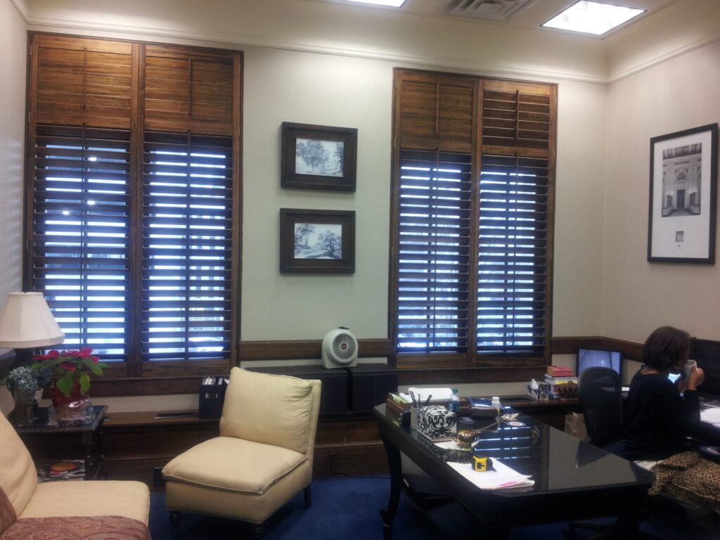 Office interior featuring plantation shutters on windows, a modern desk with office supplies, and a person seated at the desk, emphasizing the aesthetic and functional benefits of plantation shutters in a professional setting.
