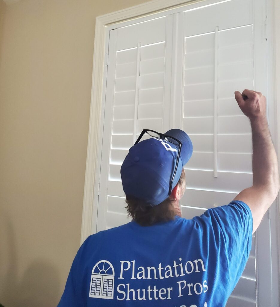 Person in blue shirt adjusting plantation shutters during installation, showcasing Plantation Shutter Pros branding.