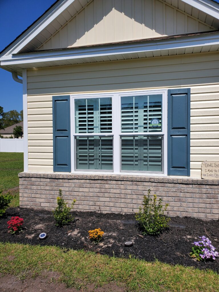 Plantation shutters on a residential window with blue shutters, surrounded by landscaped flower beds featuring colorful blooms and a brick foundation, showcasing window treatment options for home enhancement.