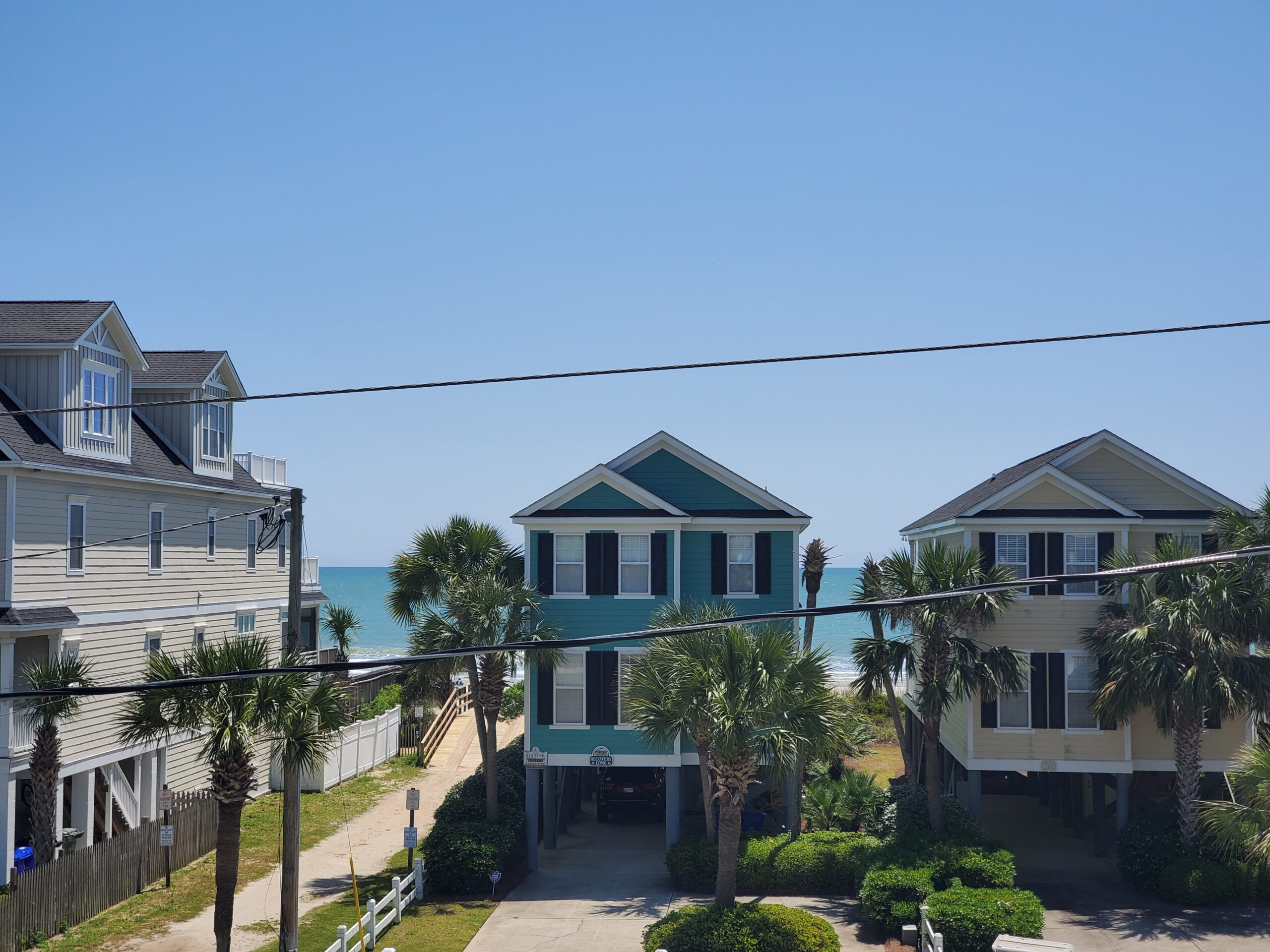 Caswell Beach, NC coastal view featuring two homes with palm trees and ocean backdrop, highlighting potential locations for plantation shutters and window treatments.
