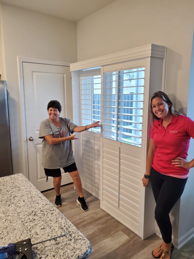Two women smiling and posing next to newly installed plantation shutters in a modern home setting, showcasing quality window treatments by Plantation Shutter Pros.