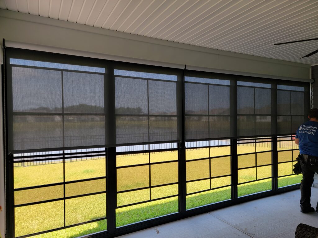 Exterior solar shades installed on a porch, showcasing a scenic view of green grass and water, with a technician from Plantation Shutter Pros adjusting the shades.