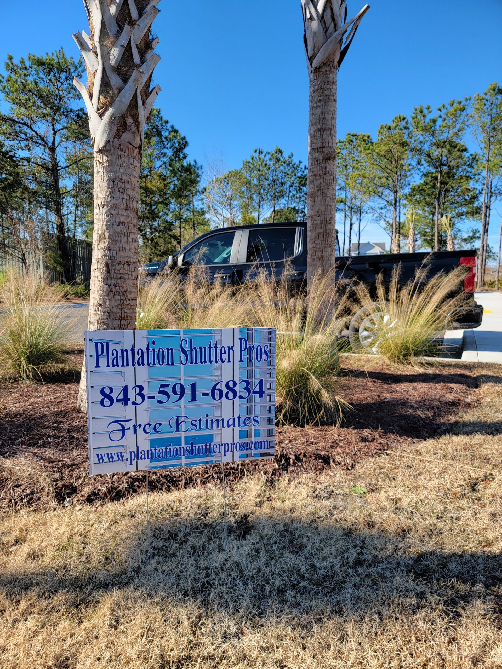 Plantation Shutter Pros sign displaying contact information and free estimates in a landscaped area with palm trees and a black truck in the background.