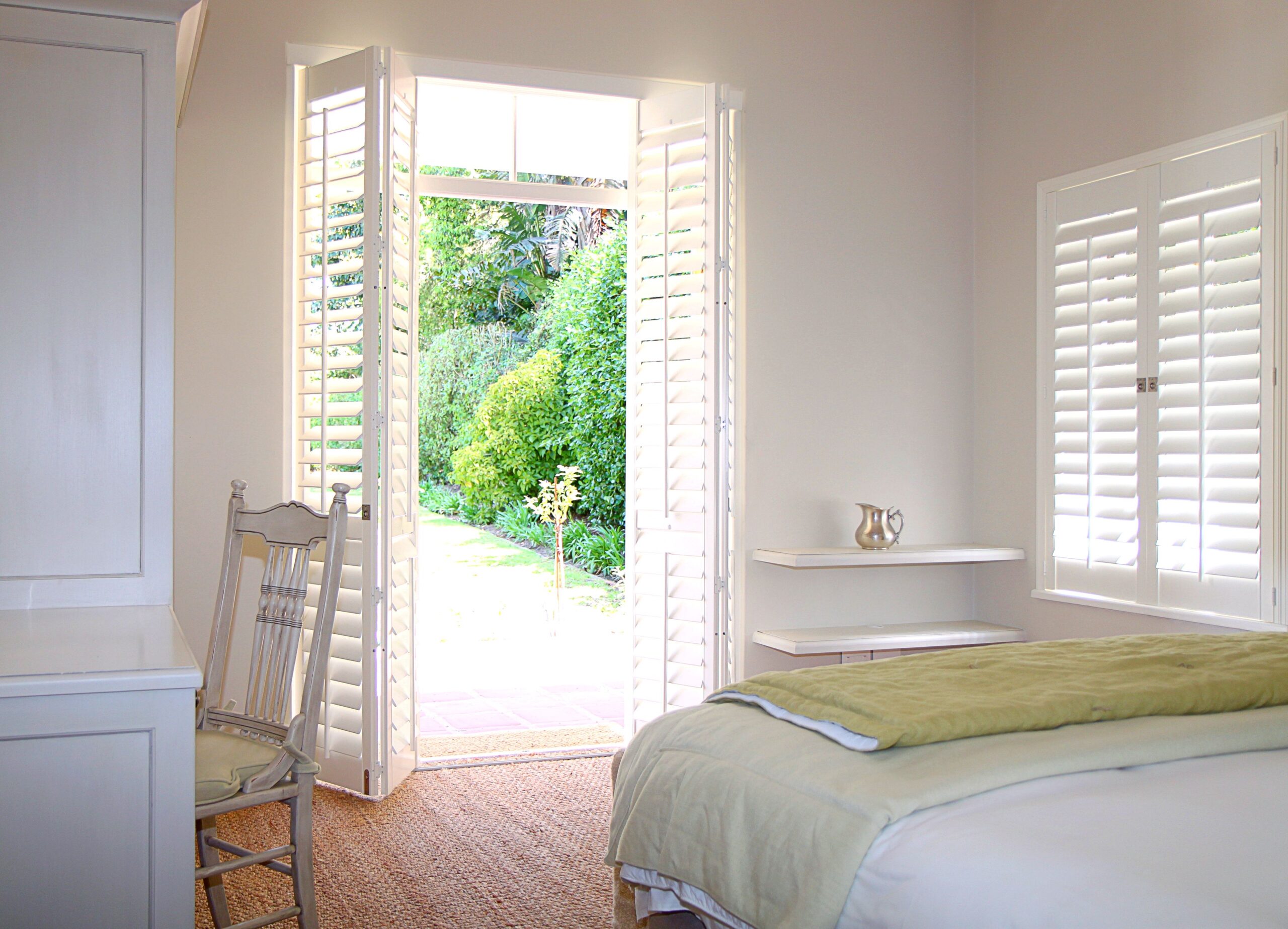 Bright bedroom interior featuring open plantation shutters, a view of lush greenery outside, and a neatly made bed with a green throw blanket, emphasizing energy-efficient home design.