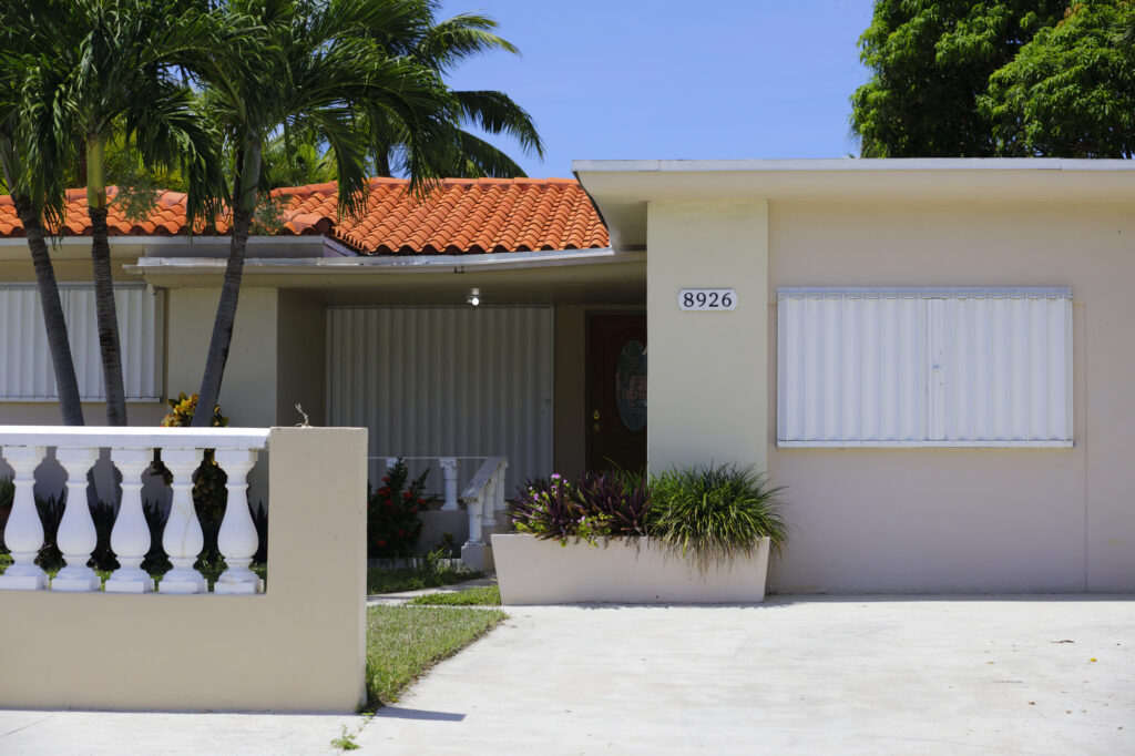 Residential home exterior with tropical landscaping, featuring decorative white railing, palm trees, and a distinctive orange tiled roof, showcasing hurricane shutters on windows, emphasizing outdoor solutions for enhanced home protection.