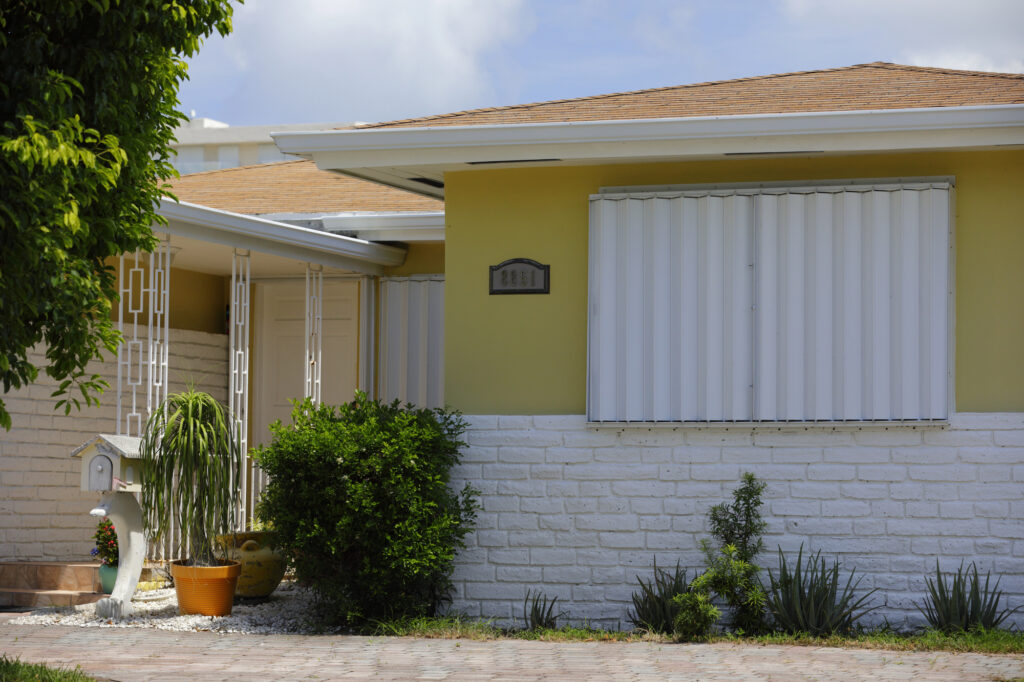 Exterior view of a residential home featuring white accordion shutters, a yellow wall, and landscaped greenery, showcasing window treatment solutions for enhanced home aesthetics.
