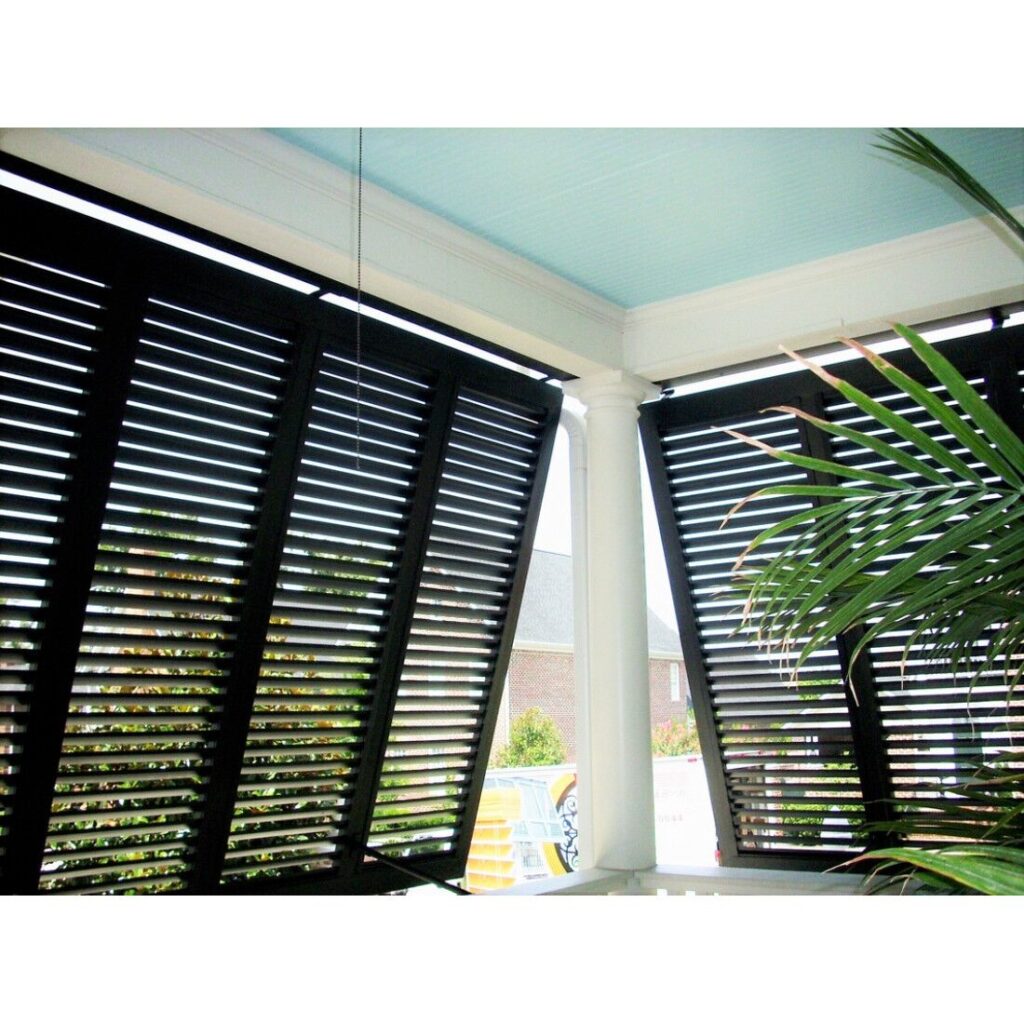 Bahama hurricane shutters with articulated blades, showcasing a black finish, installed on a porch with a blue ceiling and visible greenery.