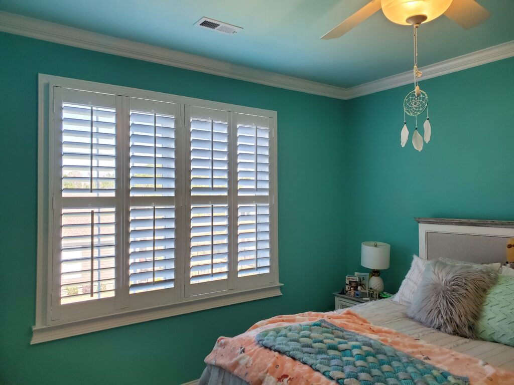 Bright bedroom featuring white plantation shutters on a turquoise wall, cozy bedding with pastel colors, and a ceiling fan, showcasing a stylish window treatment option.