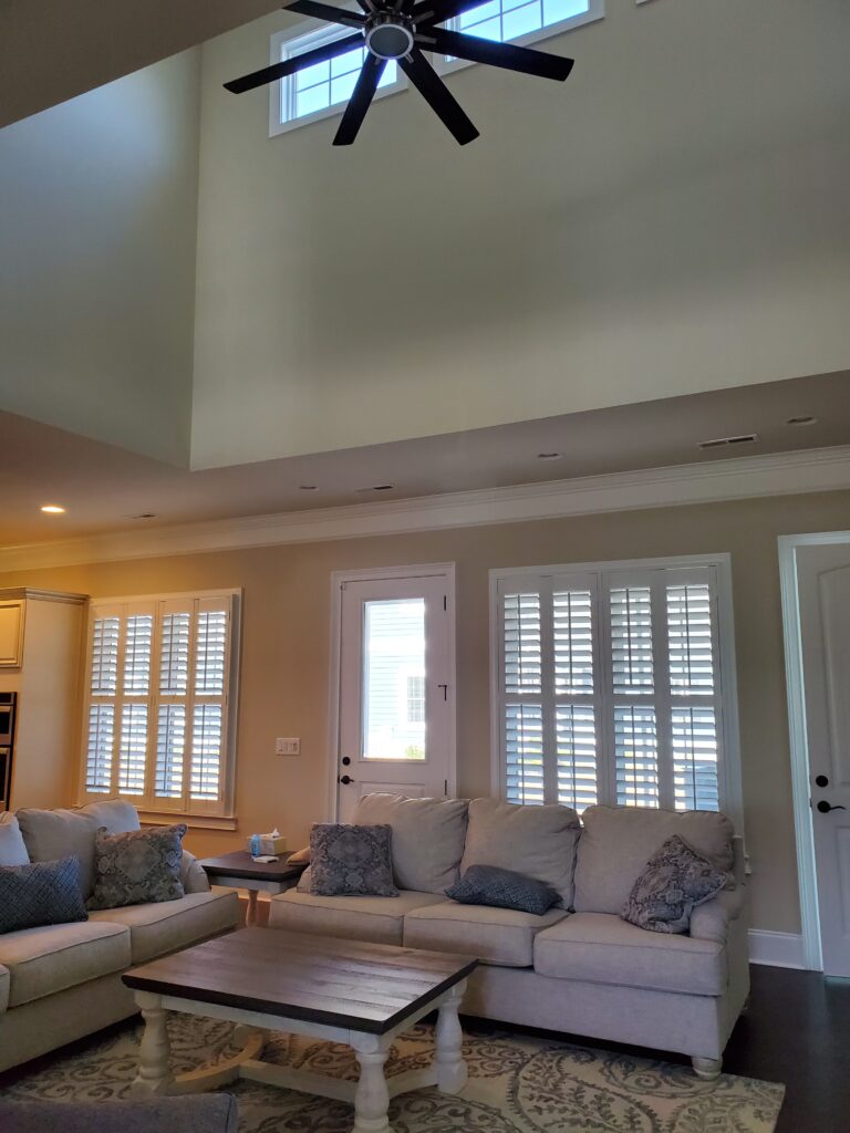Living room featuring plantation shutters on windows, a ceiling fan, beige walls, and a cozy seating arrangement with decorative pillows and a wooden coffee table.