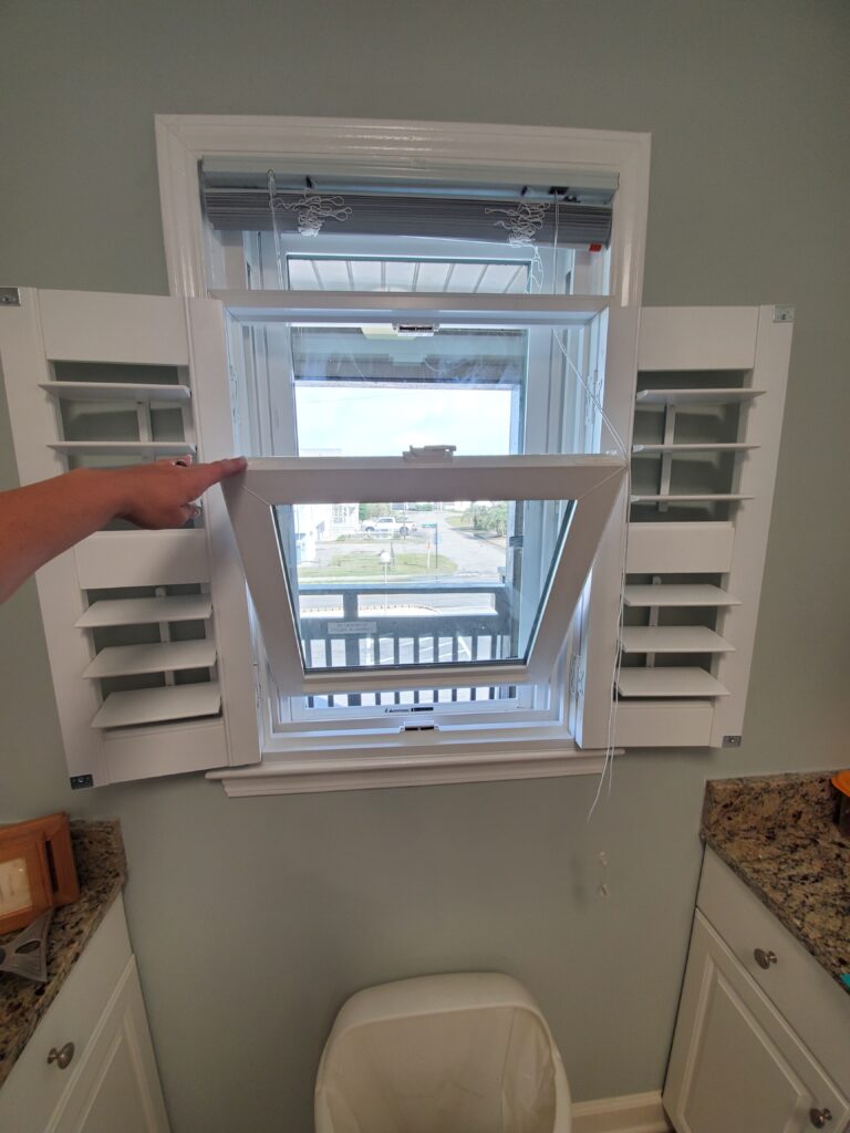 Hand opening hybrid plantation shutters to reveal a window, showcasing a view outside, in a kitchen setting with granite countertops and a trash bin.