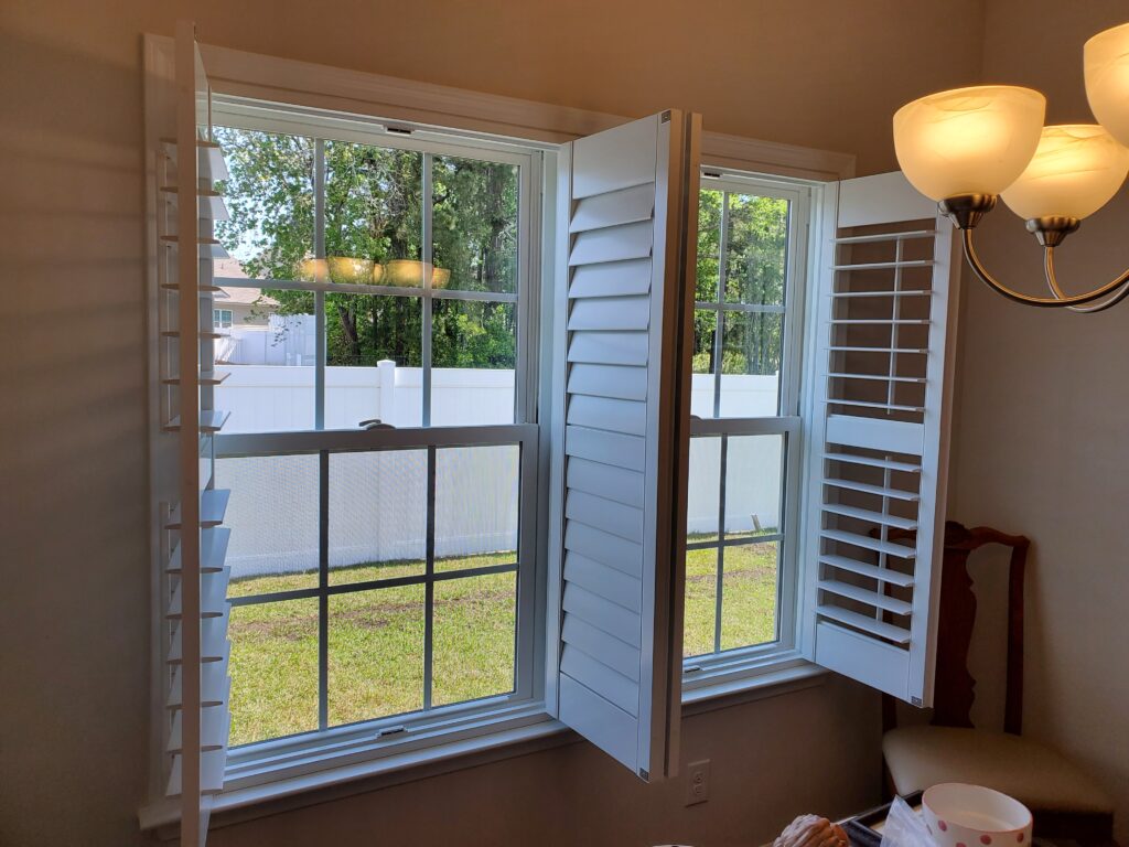 White plantation shutters opened in a bright room, showcasing a view of a green yard and white fence, highlighting window treatment options for residential spaces.