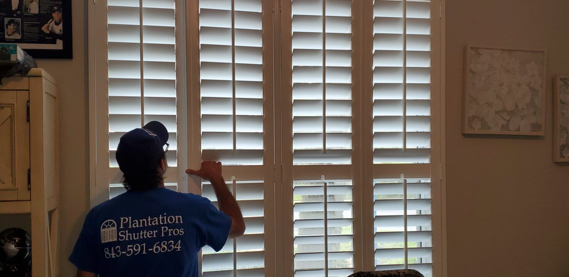 Man adjusting plantation shutters in a home, wearing a blue shirt with "Plantation Shutter Pros" logo, highlighting window treatment installation.