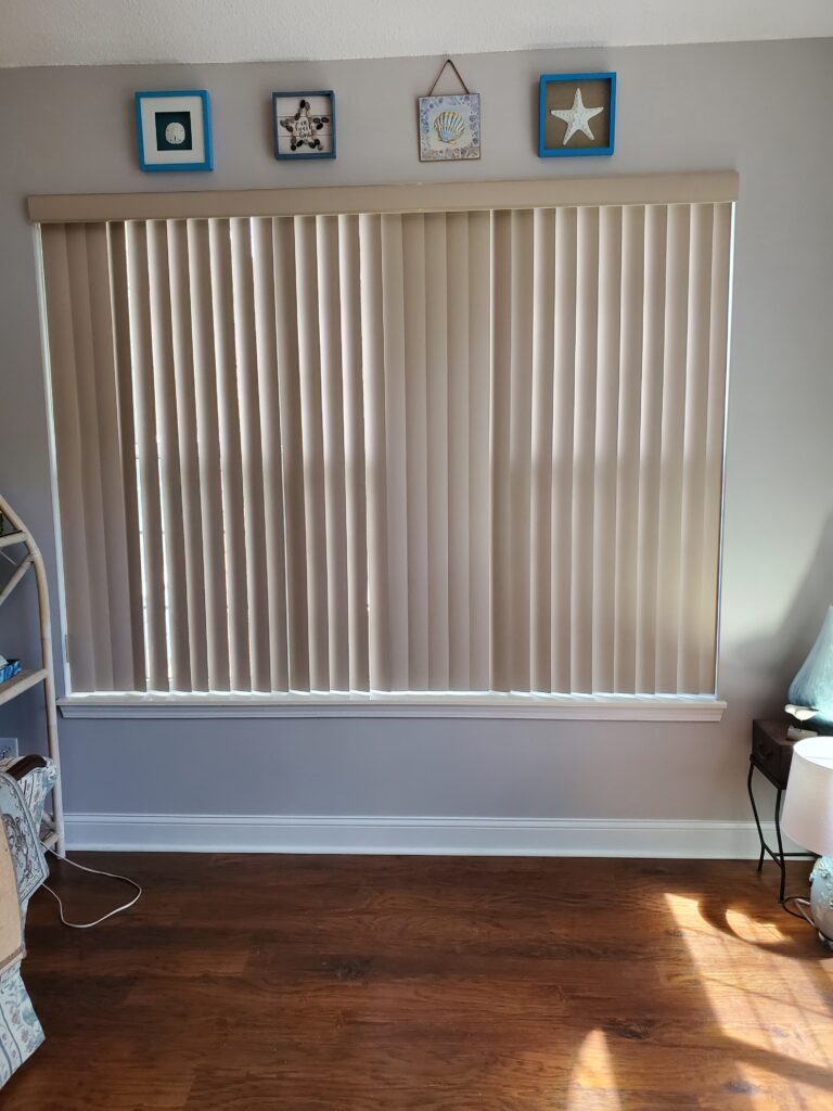 Vertical beige blinds covering a window in a cozy room, with decorative wall art above and natural light illuminating the wooden floor.