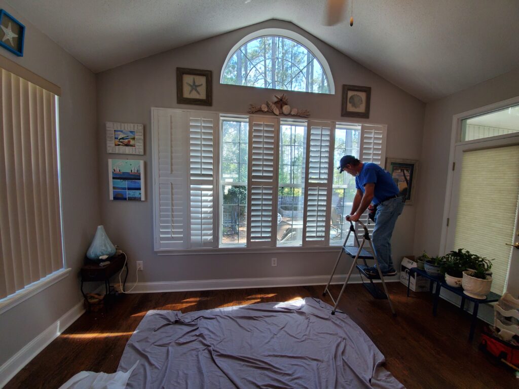 Man installing plantation shutters in a sunlit room with wooden floors, featuring decorative wall art and a cozy atmosphere.