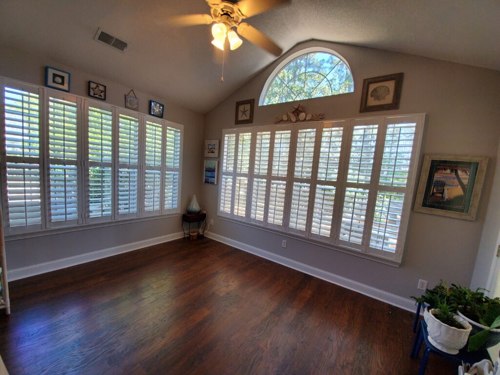 Bright room featuring plantation shutters on large windows, showcasing natural light and a cozy interior with wooden flooring and decorative elements.