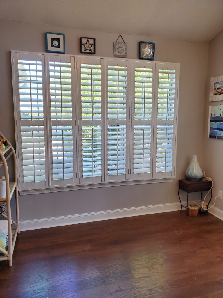 Interior view of a room featuring white plantation shutters on a large window, framed by decorative wall art and a wooden floor, showcasing a stylish window treatment option for home aesthetics.