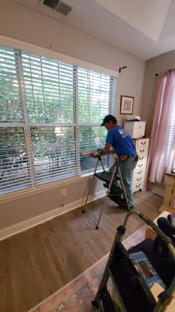 Technician installing faux wood blinds in a residential room with natural light and greenery visible outside the window.