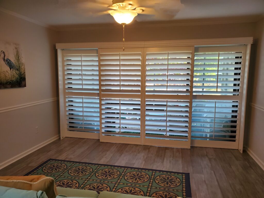 Interior view of a room featuring white plantation shutters on a large window, with a decorative rug and wall art, showcasing a stylish window treatment option for enhancing home aesthetics.