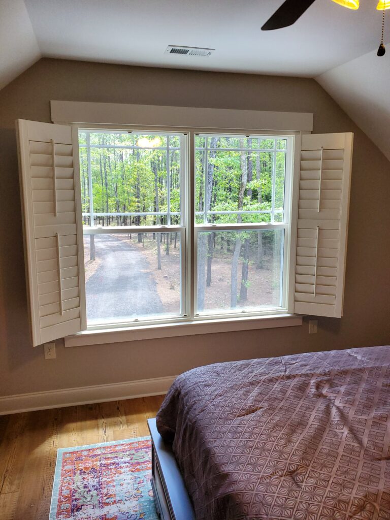 Window featuring hybrid plantation shutters in a cozy bedroom, overlooking a wooded area and a gravel path, enhancing natural light and aesthetics.
