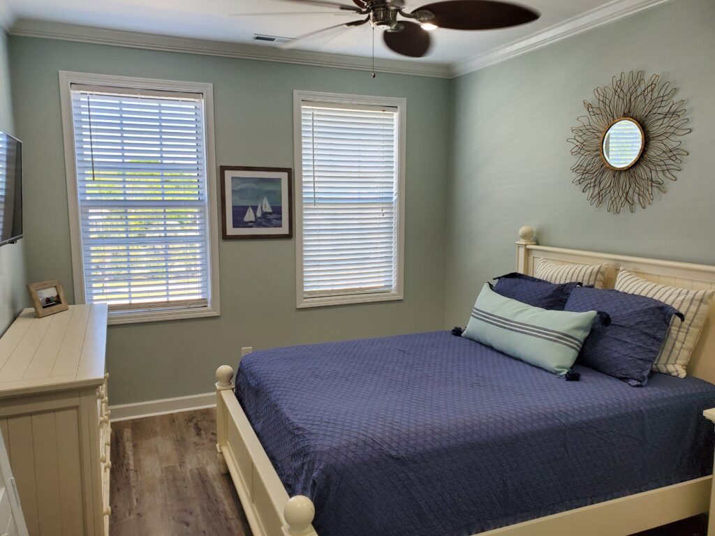 Bedroom featuring plantation shutters on windows, a white bed with blue bedding, decorative pillows, and a wall-mounted television, enhancing a coastal aesthetic.