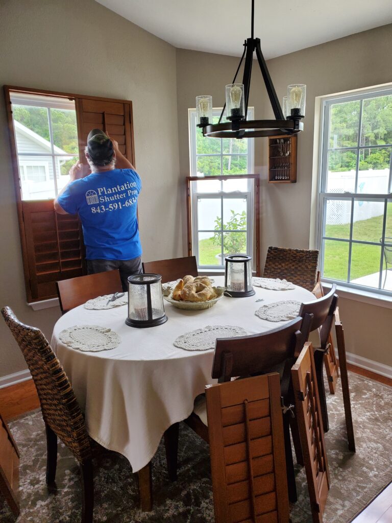 Man in blue Plantation Shutter Pros shirt installing plantation shutters in a dining area with a round table, woven chairs, and natural light from windows.