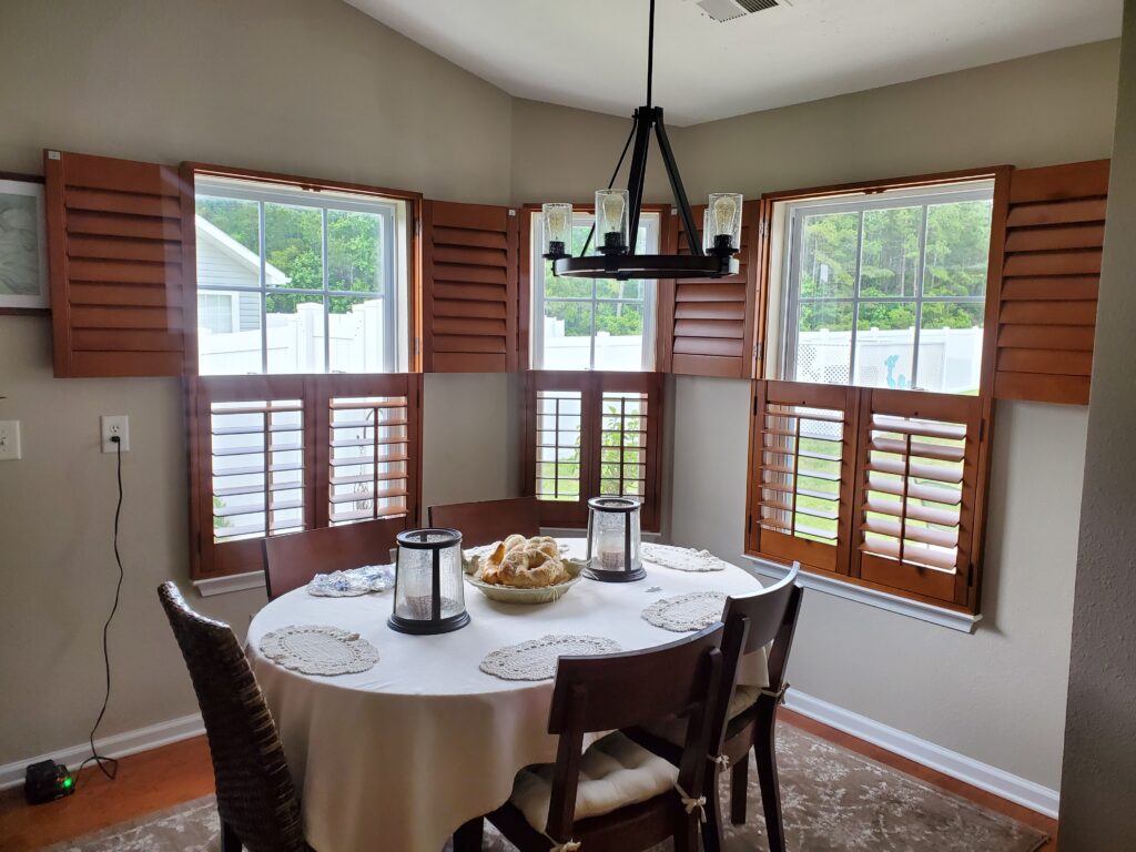 Dining area featuring wooden plantation shutters, round table set with decorative tableware, and a chandelier, enhancing the aesthetic of window treatments in a residential space.