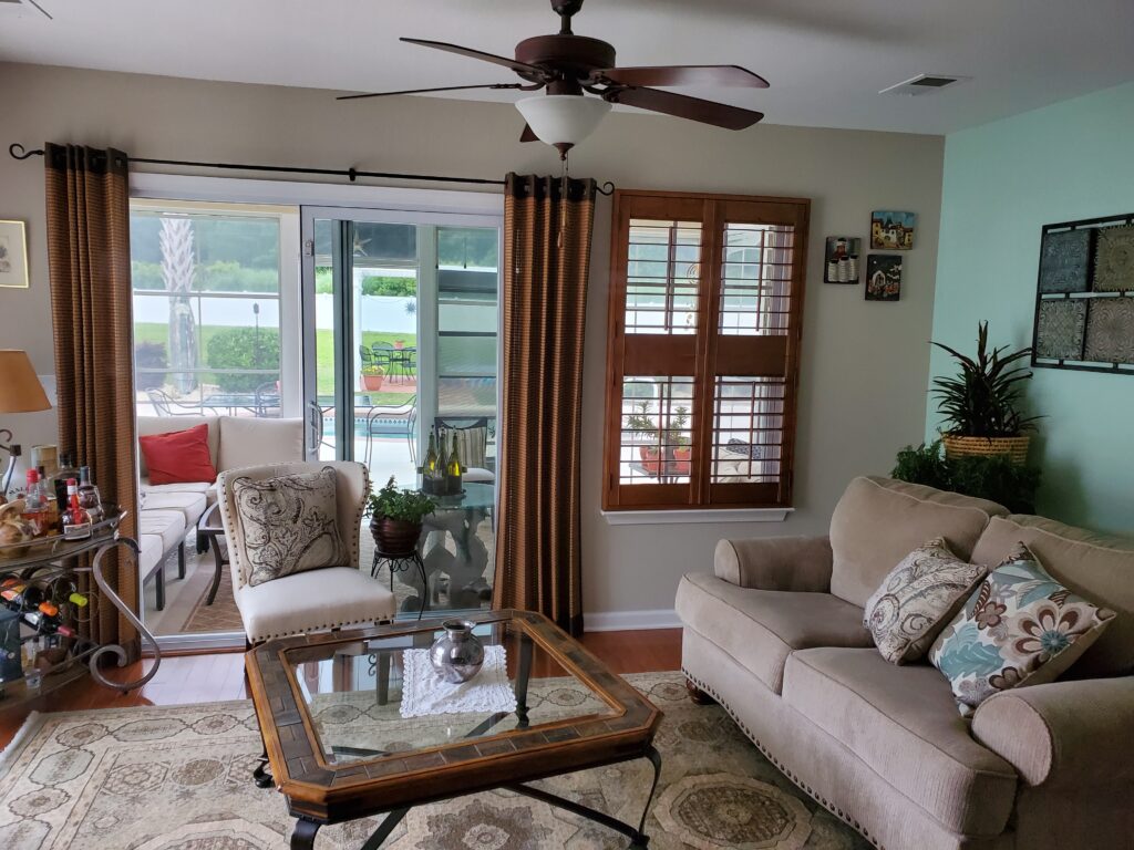 Living room featuring plantation shutters, comfortable sofa with decorative pillows, and a coffee table, showcasing a bright, inviting space with outdoor views.