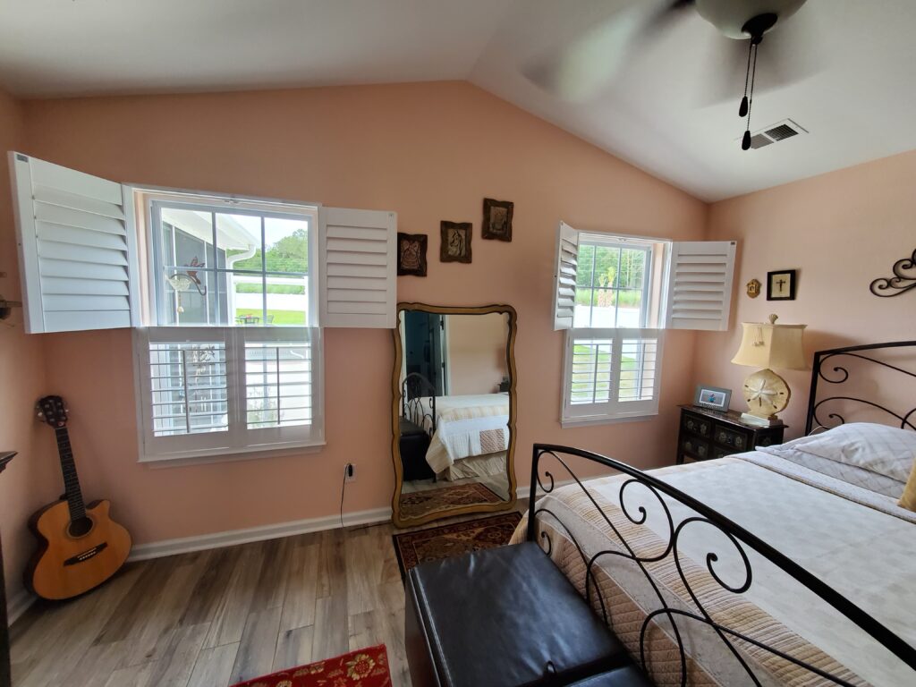 Bedroom featuring hybrid plantation shutters on windows, a decorative mirror, acoustic guitar, and cozy furnishings, showcasing stylish window treatments for enhancing home aesthetics.