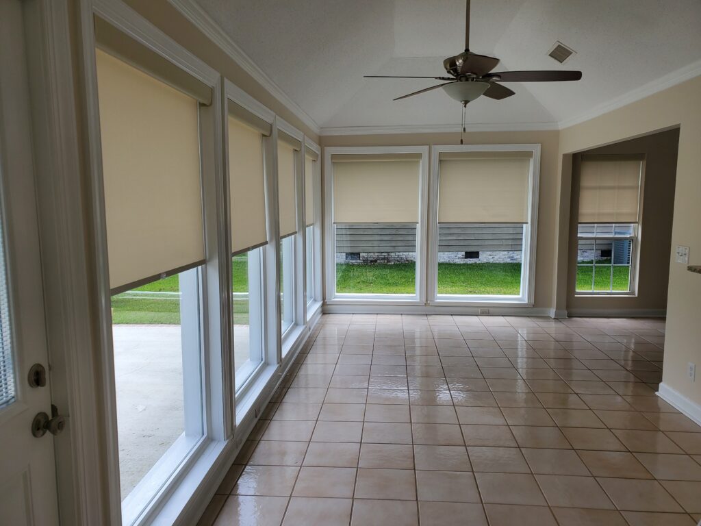 Interior view of a sunlit room featuring beige roller shades on large windows, tiled flooring, and a ceiling fan, showcasing a modern window treatment option for home aesthetics.