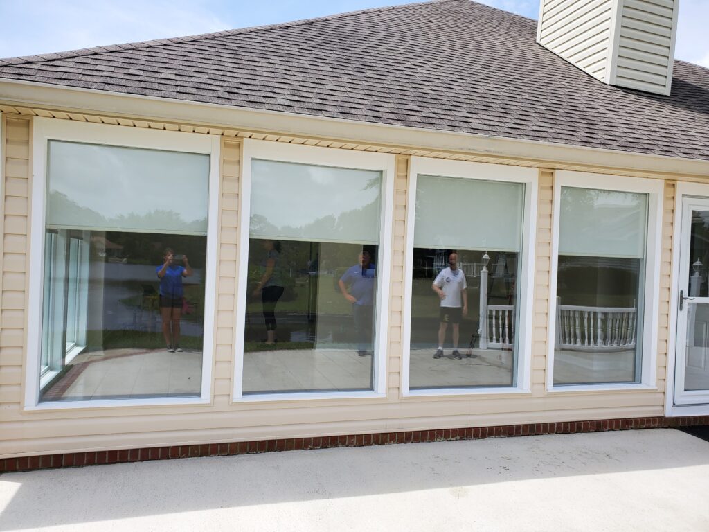 Modern home exterior featuring large windows with sheer shades, showcasing reflections of people outside, complementing the gallery of window treatments by Plantation Shutter Pros.