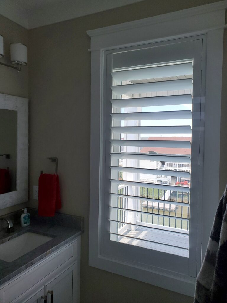 Bathroom window featuring hybrid plantation shutters, allowing natural light, with a view of a deck and water, complemented by a modern sink and red towel.