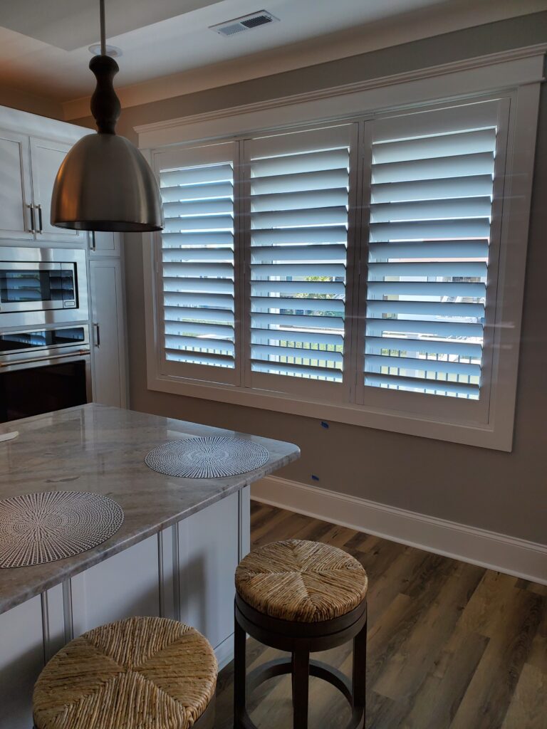 Modern kitchen featuring plantation shutters on a large window, with a marble countertop and woven bar stools, enhancing the aesthetic appeal of the space.
