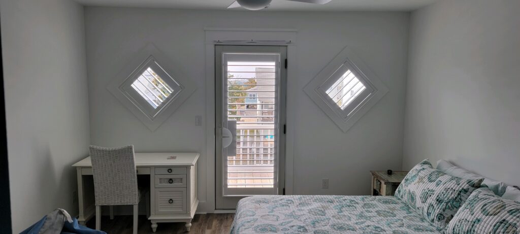 Bright bedroom featuring plantation shutters on diamond-shaped windows, a white desk with a wicker chair, and a bed adorned with a patterned quilt, enhancing the aesthetic of the space.