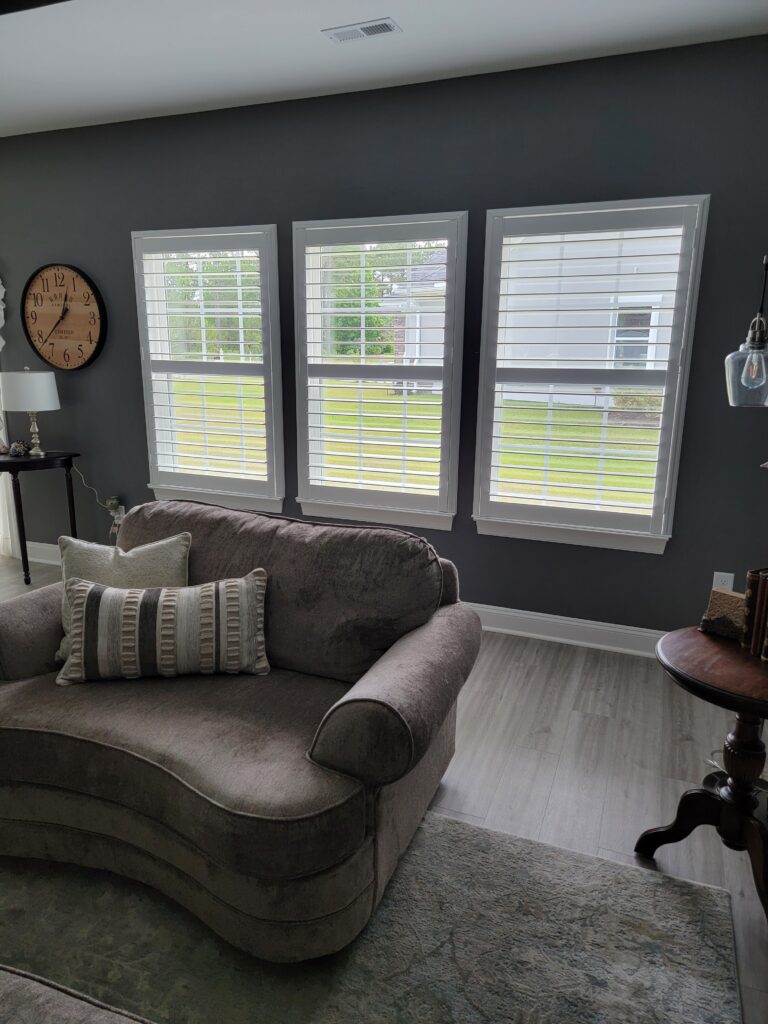 Cozy living room featuring three plantation shuttered windows, a plush curved sofa with decorative pillows, and a stylish clock on the wall, enhancing the aesthetic appeal of the space.