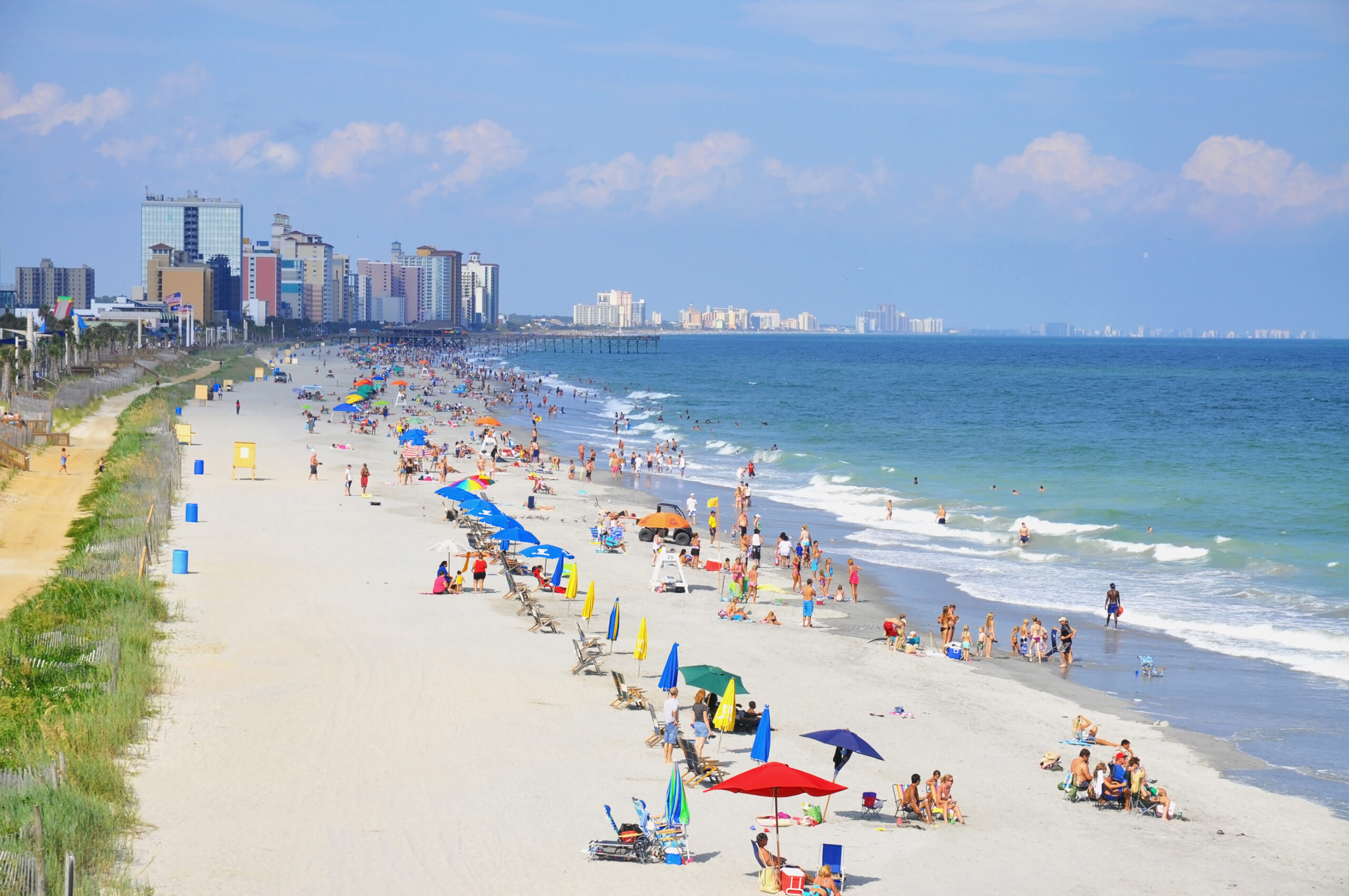 Crowded beach scene in Myrtle Beach, South Carolina, featuring colorful umbrellas, sunbathers, and a skyline of high-rise buildings along the coastline.