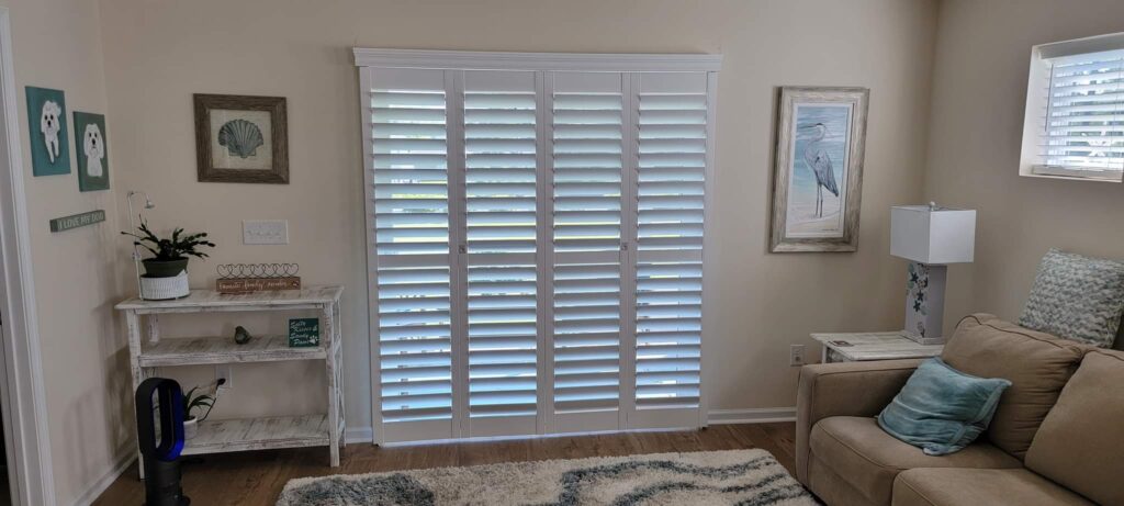 Interior living space featuring white plantation shutters, beige walls, a cozy couch with decorative pillows, a side table with a lamp, and decorative wall art, emphasizing window treatments for home enhancement.